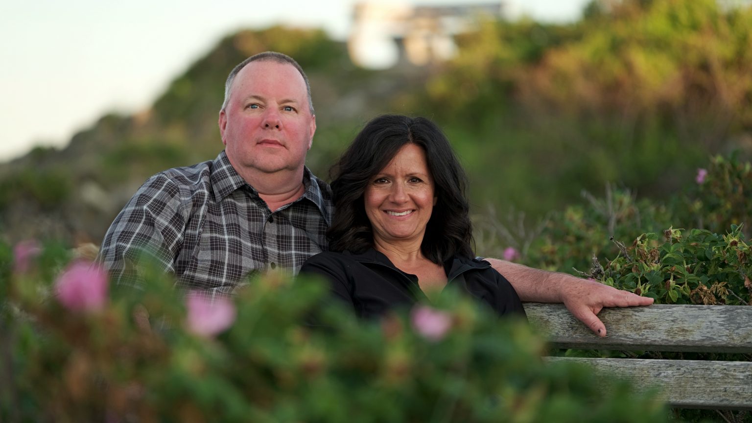 Paul Hackett and his wife seated together on a wooden bench among coastal wild roses